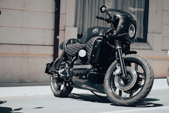 A sleek black motorcycle with a quilted seat and a large front headlight is parked in front of a building with beige brick walls and a curtained window. The motorcycle has distinctive branding, polished chrome elements, and an overall modern, custom design.