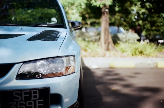 A close-up view of the front left side of a blue car with prominent headlights and a visible license plate. The background features a tree and blurred greenery, suggesting an outdoor setting.