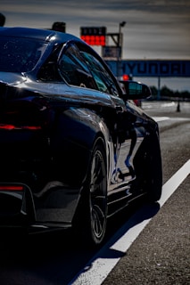 A sleek, black car is positioned on a track, ready for a race. The backdrop includes a large Hyundai banner and a digital timer displaying numbers. The lighting suggests a sunny day with clear reflections on the car's surface.