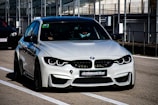 A sleek, modern BMW car is parked on a race track, positioned in the pit lane. It has a silver exterior with aggressive front grille and headlights that suggest speed and performance. A sign with 'ULTIMAVUELTA.es' is affixed to the front bumper. The background features industrial structures like barriers and railings, consistent with a motorsport environment.