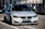 A sleek, modern BMW car is parked on a race track, positioned in the pit lane. It has a silver exterior with aggressive front grille and headlights that suggest speed and performance. A sign with 'ULTIMAVUELTA.es' is affixed to the front bumper. The background features industrial structures like barriers and railings, consistent with a motorsport environment.