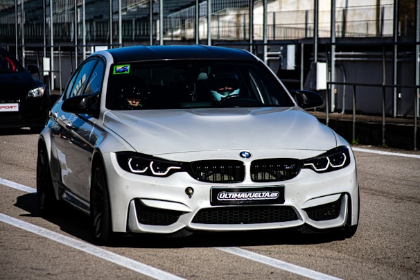 A sleek, modern BMW car is parked on a race track, positioned in the pit lane. It has a silver exterior with aggressive front grille and headlights that suggest speed and performance. A sign with 'ULTIMAVUELTA.es' is affixed to the front bumper. The background features industrial structures like barriers and railings, consistent with a motorsport environment.