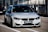 A sleek, modern BMW car is parked on a race track, positioned in the pit lane. It has a silver exterior with aggressive front grille and headlights that suggest speed and performance. A sign with 'ULTIMAVUELTA.es' is affixed to the front bumper. The background features industrial structures like barriers and railings, consistent with a motorsport environment.