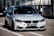 A sleek, modern BMW car is parked on a race track, positioned in the pit lane. It has a silver exterior with aggressive front grille and headlights that suggest speed and performance. A sign with 'ULTIMAVUELTA.es' is affixed to the front bumper. The background features industrial structures like barriers and railings, consistent with a motorsport environment.