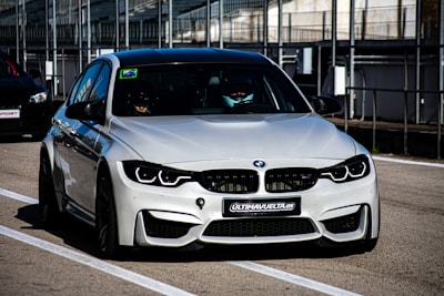 A sleek, modern BMW car is parked on a race track, positioned in the pit lane. It has a silver exterior with aggressive front grille and headlights that suggest speed and performance. A sign with 'ULTIMAVUELTA.es' is affixed to the front bumper. The background features industrial structures like barriers and railings, consistent with a motorsport environment.