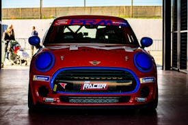 A red and blue racing car is parked inside a garage, with a focus on its front view. The car features prominent branding and logos, including a Mini emblem and various racing decals. The bright blue headlights and details contrast with the red body. In the background, some people are partially visible near the entrance, with a stroller suggesting a family atmosphere outside.