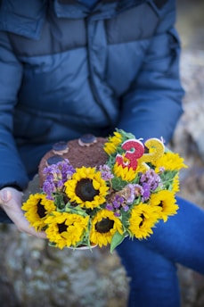 A person in a blue jacket is holding a bouquet of sunflowers and purple flowers alongside a chocolate cake. The cake has two glittery candles shaped as the number 3.