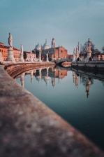 A serene architectural scene features statues lining a canal, with a historical building in the background. The reflections in the calm water create a mirror image effect, enhancing the symmetrical beauty of the setting.