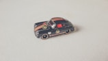 A small, detailed black toy car with gold and red stripes, placed on a light surface. It features branding and logo details that mimic a classic racing design.