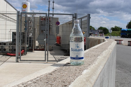A clear glass bottle labeled 'Bismarck' is placed on a concrete railing in an industrial area. Behind the bottle, there is a fenced-off section with industrial equipment, pipes, and a sign indicating the presence of a Shell gas facility. The sky is partly cloudy, and a blue truck is visible in the background.