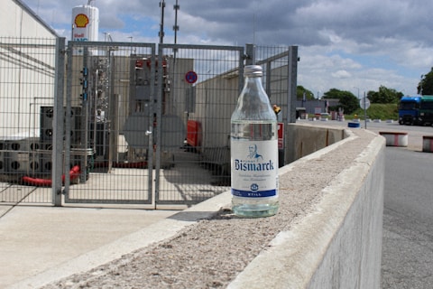 A clear glass bottle labeled 'Bismarck' is placed on a concrete railing in an industrial area. Behind the bottle, there is a fenced-off section with industrial equipment, pipes, and a sign indicating the presence of a Shell gas facility. The sky is partly cloudy, and a blue truck is visible in the background.