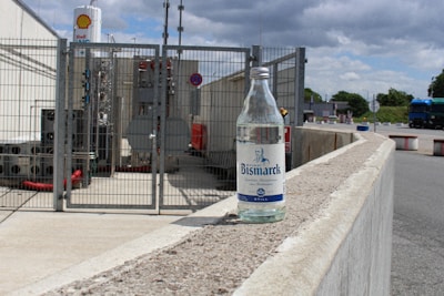 A clear glass bottle labeled 'Bismarck' is placed on a concrete railing in an industrial area. Behind the bottle, there is a fenced-off section with industrial equipment, pipes, and a sign indicating the presence of a Shell gas facility. The sky is partly cloudy, and a blue truck is visible in the background.