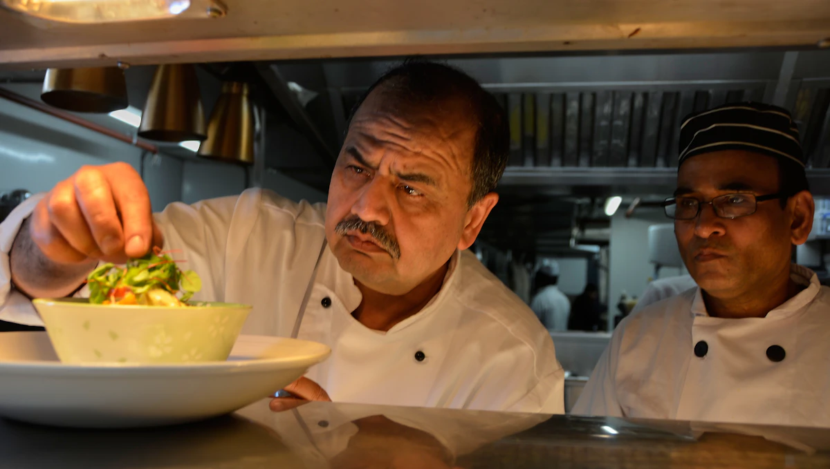 Two chefs at a restaurant pass examining a finished bowl of salad, one adding a garnish