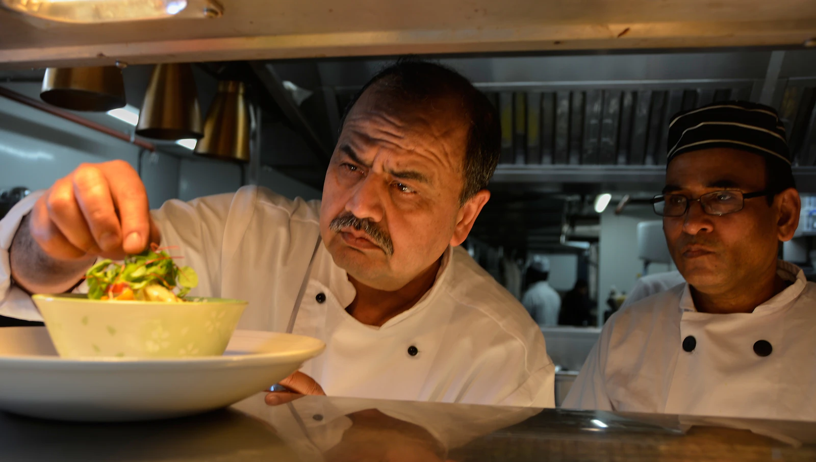 Senior chef in whites tasting a dish at the pass while a trainee looks on