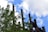 Tall industrial smokestacks rise against a backdrop of a blue sky dotted with white clouds. In the foreground, lush green tree branches provide a stark contrast to the rusty brown of the industrial structures.