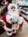 A decorative Santa Claus figure is dressed in a classic red and white outfit with a fluffy white beard and glasses. The figure holds a straw ornament and a sack filled with pinecones and greenery. Surrounding the figure is a festive store setting with holiday decorations and blurred shelves in the background.