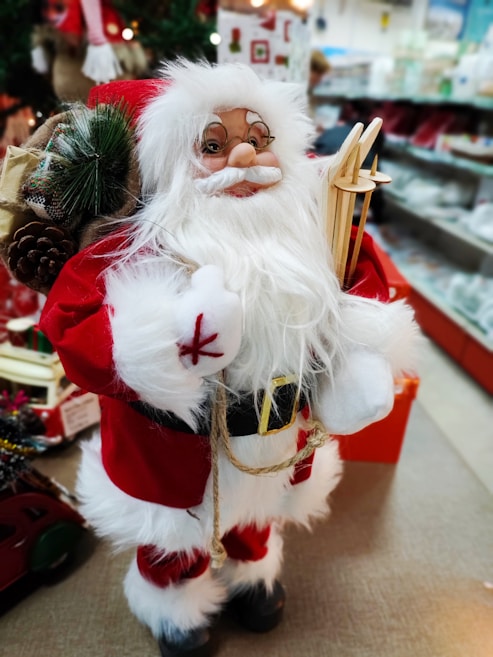 A decorative Santa Claus figure is dressed in a classic red and white outfit with a fluffy white beard and glasses. The figure holds a straw ornament and a sack filled with pinecones and greenery. Surrounding the figure is a festive store setting with holiday decorations and blurred shelves in the background.