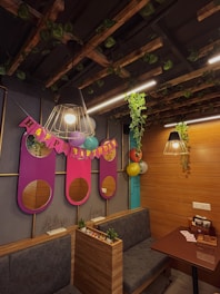 A cozy room decorated for a birthday celebration with wooden paneling and hanging lights. Vines decorate the ceiling and a 'Happy Birthday' banner is prominently displayed with colorful balloons. There are plush seating arrangements and small potted plants add a touch of greenery.
