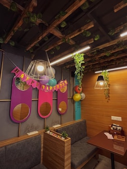 A cozy room decorated for a birthday celebration with wooden paneling and hanging lights. Vines decorate the ceiling and a 'Happy Birthday' banner is prominently displayed with colorful balloons. There are plush seating arrangements and small potted plants add a touch of greenery.