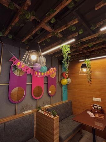 A cozy room decorated for a birthday celebration with wooden paneling and hanging lights. Vines decorate the ceiling and a 'Happy Birthday' banner is prominently displayed with colorful balloons. There are plush seating arrangements and small potted plants add a touch of greenery.