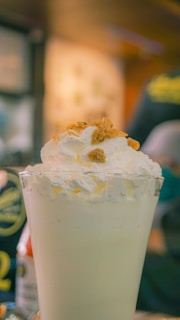 A tall glass filled with a creamy milkshake topped with whipped cream and small pieces of granola. The background is blurred, suggesting a café or restaurant setting.