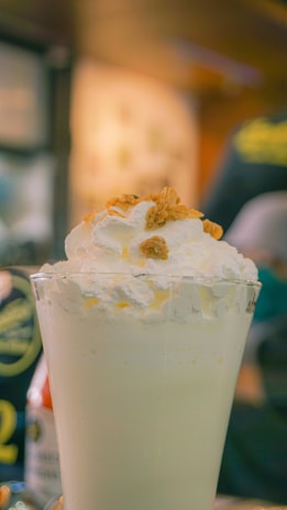 A tall glass filled with a creamy milkshake topped with whipped cream and small pieces of granola. The background is blurred, suggesting a café or restaurant setting.