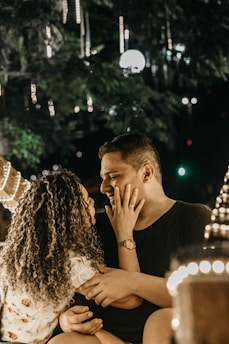 A couple sitting closely in a softly lit outdoor setting, with strings of lights hanging around them. The woman gently touches the man's face while looking into his eyes. The surroundings suggest a romantic, intimate atmosphere.