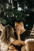 A couple sitting closely in a softly lit outdoor setting, with strings of lights hanging around them. The woman gently touches the man's face while looking into his eyes. The surroundings suggest a romantic, intimate atmosphere.