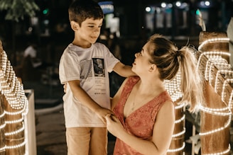 A young boy and a woman are interacting in a warmly lit outdoor setting. The boy is wearing a white t-shirt with graphics and light-colored shorts, while the woman is in a pink sleeveless top. They are surrounded by a railing decorated with glowing string lights, creating a cozy atmosphere in the evening or night-time ambiance.