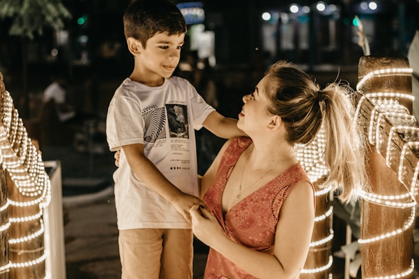 A young boy and a woman are interacting in a warmly lit outdoor setting. The boy is wearing a white t-shirt with graphics and light-colored shorts, while the woman is in a pink sleeveless top. They are surrounded by a railing decorated with glowing string lights, creating a cozy atmosphere in the evening or night-time ambiance.