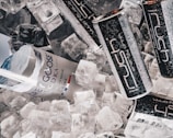 A mix of ice cubes surrounds a Grey Goose vodka bottle and several Rush energy drink cans. The items are closely arranged, giving a refreshing and chilled appearance. The surface of the cans and bottle shows condensation, emphasizing the cold atmosphere.