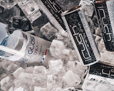 A mix of ice cubes surrounds a Grey Goose vodka bottle and several Rush energy drink cans. The items are closely arranged, giving a refreshing and chilled appearance. The surface of the cans and bottle shows condensation, emphasizing the cold atmosphere.
