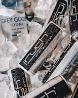 Cans of energy drink and a bottle of vodka are placed among ice cubes, with water droplets visible on the surfaces, suggesting they are chilled.