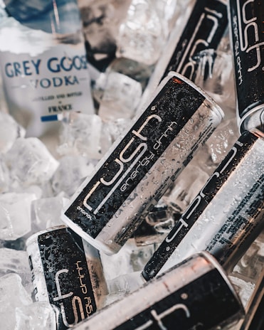 Cans of energy drink and a bottle of vodka are placed among ice cubes, with water droplets visible on the surfaces, suggesting they are chilled.