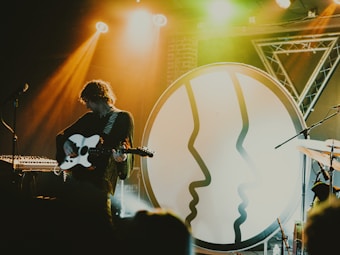 A musician is playing an electric guitar on stage with dramatic lighting and a large circular backdrop featuring abstract face profiles. The setting includes stage equipment such as microphones and keyboards, with warm yellow and green lights creating a vibrant atmosphere.