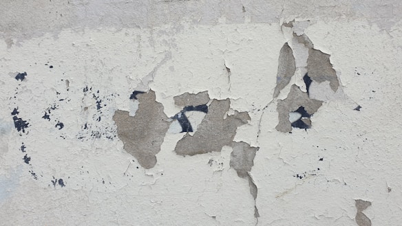 Peeling and cracked plaster on a wall, revealing darker patches underneath. The surface shows various layers of wear over time, creating an aged and weathered appearance.
