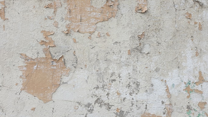 A weathered, peeling wall with patches of exposed plaster and paint. The surface shows signs of aging with areas where the top layer of paint has chipped off, revealing an underlying layer. The wall has a rough texture and varying shades from beige to light brown.