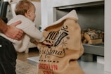 A young child reaching into a burlap sack with festive writing, possibly Christmas-themed, in a cozy indoor setting. An adult hand gently supports the child, creating a warm, familial atmosphere. Firewood is stacked in the background, adding to the cozy vibe.