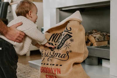 A young child reaching into a burlap sack with festive writing, possibly Christmas-themed, in a cozy indoor setting. An adult hand gently supports the child, creating a warm, familial atmosphere. Firewood is stacked in the background, adding to the cozy vibe.