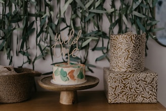 Cake with 'Wild One' topper displayed on a wooden stand. The cake features a design with green, white, and beige colors. To the right, there are two boxes with a nature-inspired pattern, and to the left, a woven basket. Green leafy decorations hang in the background.