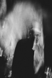A blurred, monochrome image featuring a person partially obscured by motion and lighting effects. The scene has a ghostly or abstract quality, with light streaks and shadows dominating the composition.