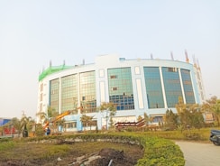 A large, modern building with blue-tinted glass windows and light-colored walls, surrounded by a landscaped area with trees and bushes. There is construction equipment, including a crane, in the foreground, indicating ongoing work.