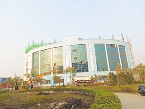 A large, modern building with blue-tinted glass windows and light-colored walls, surrounded by a landscaped area with trees and bushes. There is construction equipment, including a crane, in the foreground, indicating ongoing work.