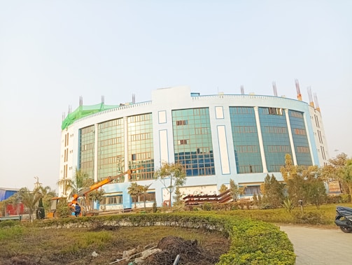 A large, modern building with blue-tinted glass windows and light-colored walls, surrounded by a landscaped area with trees and bushes. There is construction equipment, including a crane, in the foreground, indicating ongoing work.