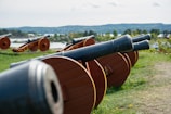 A row of historic cannons with wooden wheels is positioned on a grassy area overlooking a scenic landscape with distant hills and water. The cannons are angled upward and arranged in a line, suggesting a defensive arrangement. The setting appears to be outdoors with lush greenery and clear skies.