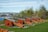 A series of old-fashioned cannons with wooden carriages painted in red with yellow detailing are lined up on a grassy area. In the background, there's a view of a marina with sailboats and a cityscape, partially obscured by trees and shrubs. The sky is clear with some light clouds.