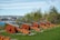 A series of old-fashioned cannons with wooden carriages painted in red with yellow detailing are lined up on a grassy area. In the background, there's a view of a marina with sailboats and a cityscape, partially obscured by trees and shrubs. The sky is clear with some light clouds.