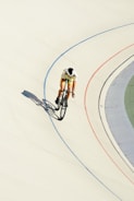A cyclist in colorful attire rides on a velodrome track, leaning into a curve with shadows cast on the light-colored surface. The track features blue and red lines marking the lanes, set against the stark contrast of the smooth white cement.