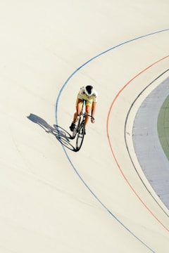 A cyclist in colorful attire rides on a velodrome track, leaning into a curve with shadows cast on the light-colored surface. The track features blue and red lines marking the lanes, set against the stark contrast of the smooth white cement.