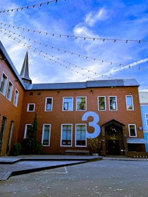 A red brick building with large windows and a prominent number three painted in white on the wall. String lights are draped across the courtyard and a tall spire is visible in the background against a blue sky with some clouds.
